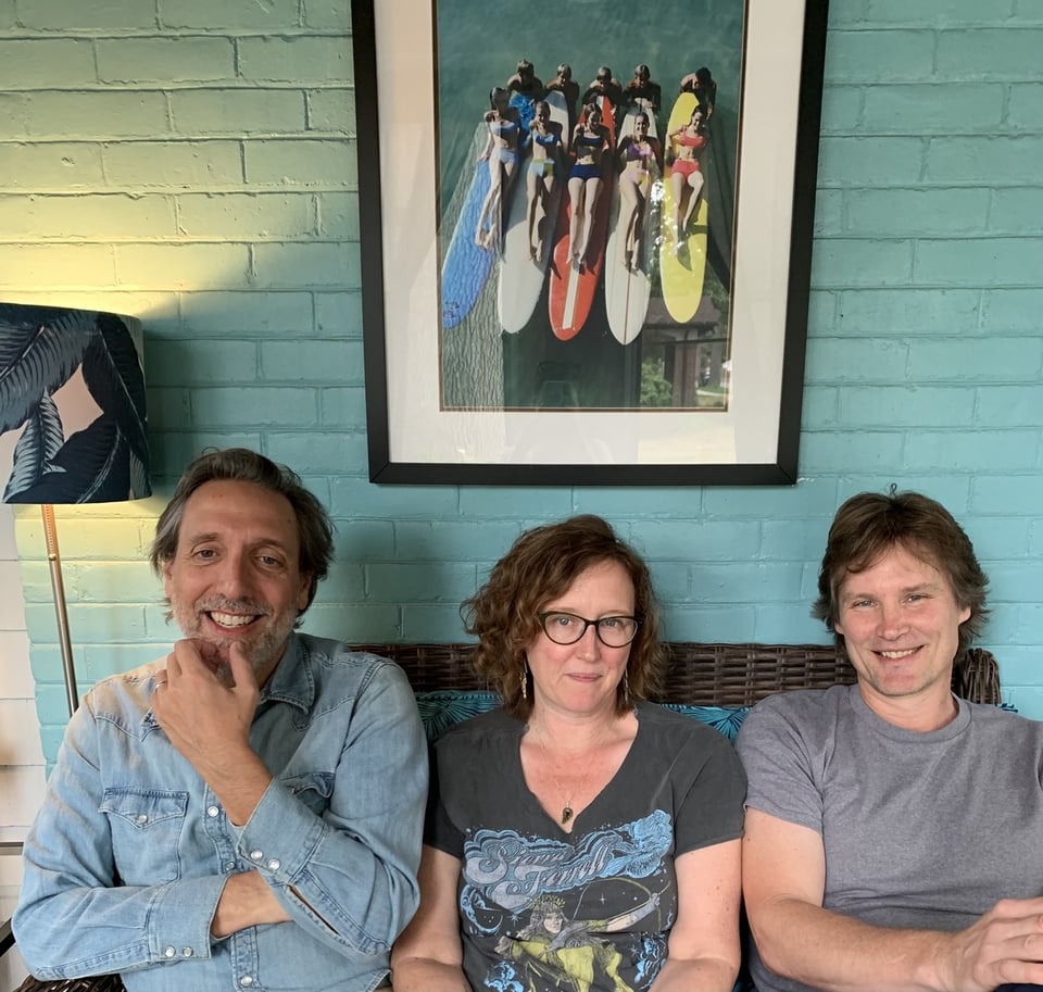 A woman with chin-length brown hair and glasses sits on a couch between two men.