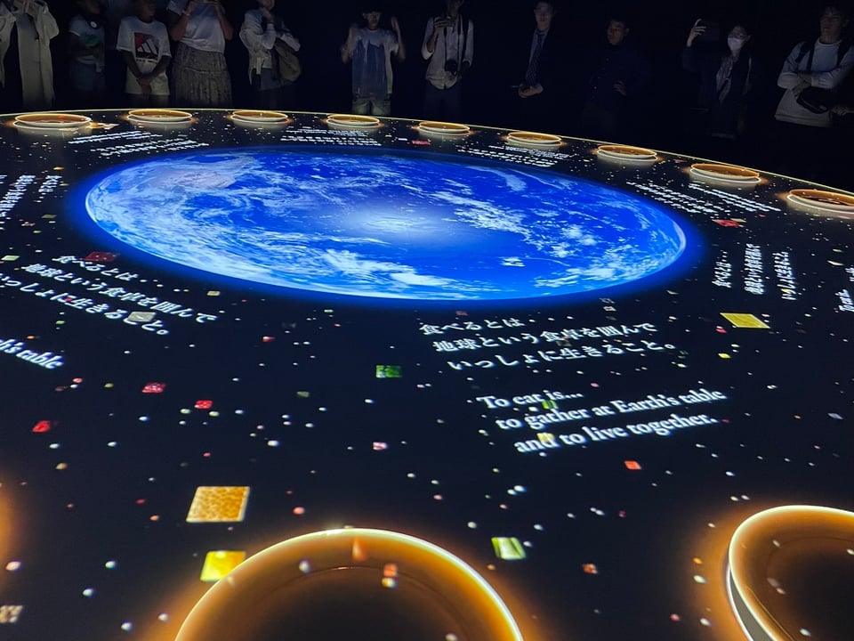 An art pieces consisting of a digital projection on a large round table set with big white plates. The earth is projected in the center, with the words "to eat is... to gather at Earth's table and to live together" in both English and Japanese.