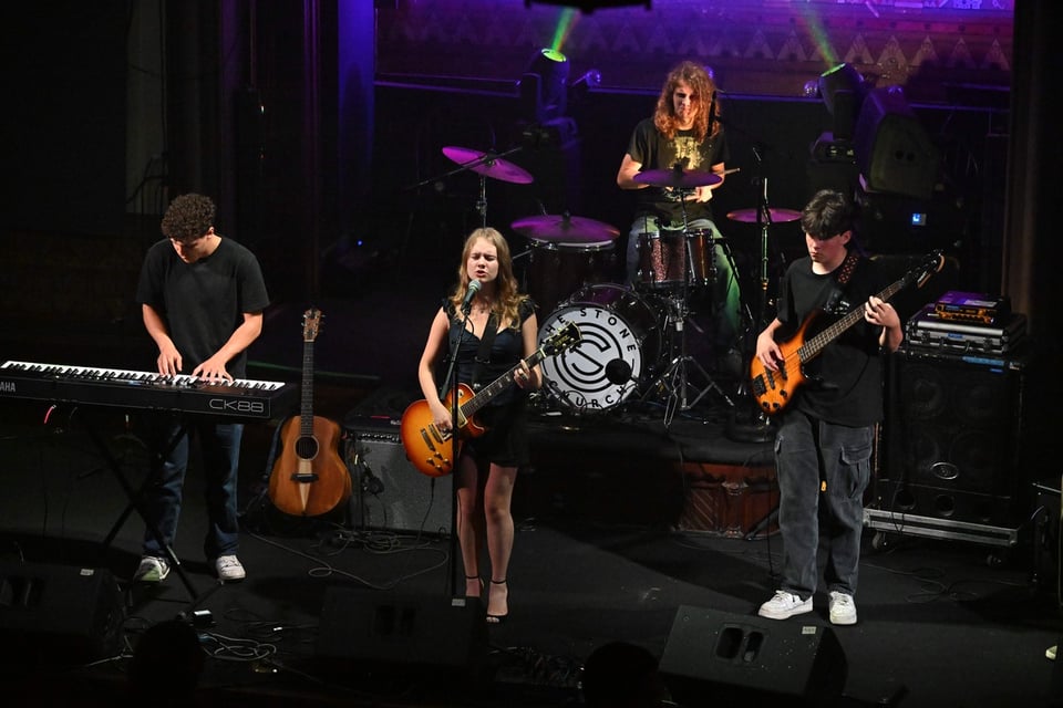A woman stands at center stage wearing a black dress with a Les Paul-style electric guitar slung over her shoulder. There’s a bassist to her left, a keyboard player to her right and a drummer behind her.