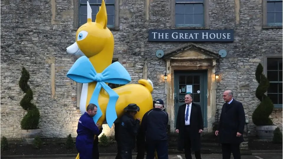 A giant statue of a stylised fawn in a huge baby blue bow is being moved by several people. It's perhaps 15 foot tall. Two older men are watching happily.