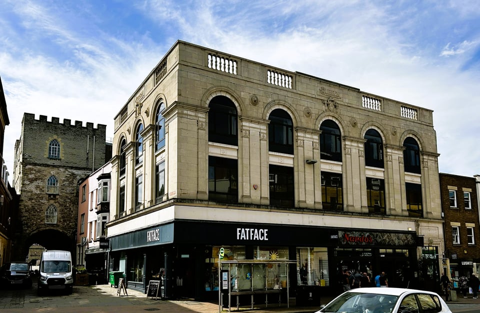 A Burtons shop, now converted into a Fatface and a food retailer. The upper floors reveal a steel frame structure, hidden behind double-height arches and stylised columns. At the top is a rather old fashioned balustrade and a plaque with the Burton's logo. To one side is the entrance to Taunton castle.