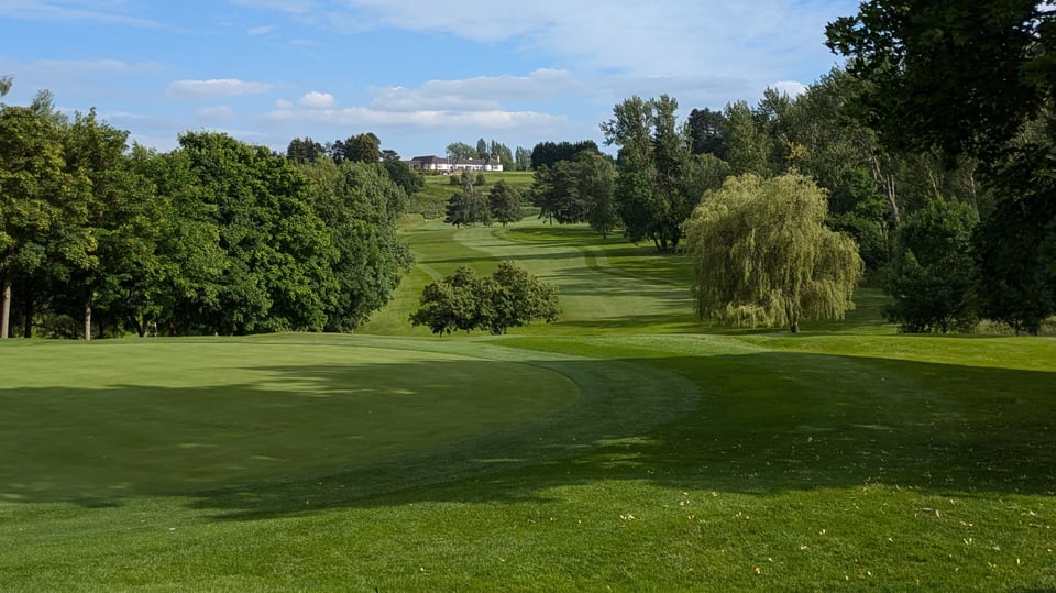 A view from the 12th green at Rushcliffe Golf Club, Nottinghamshire in summer, looking back down the tree-lined fairway and up towards the clubhouse in the far background.