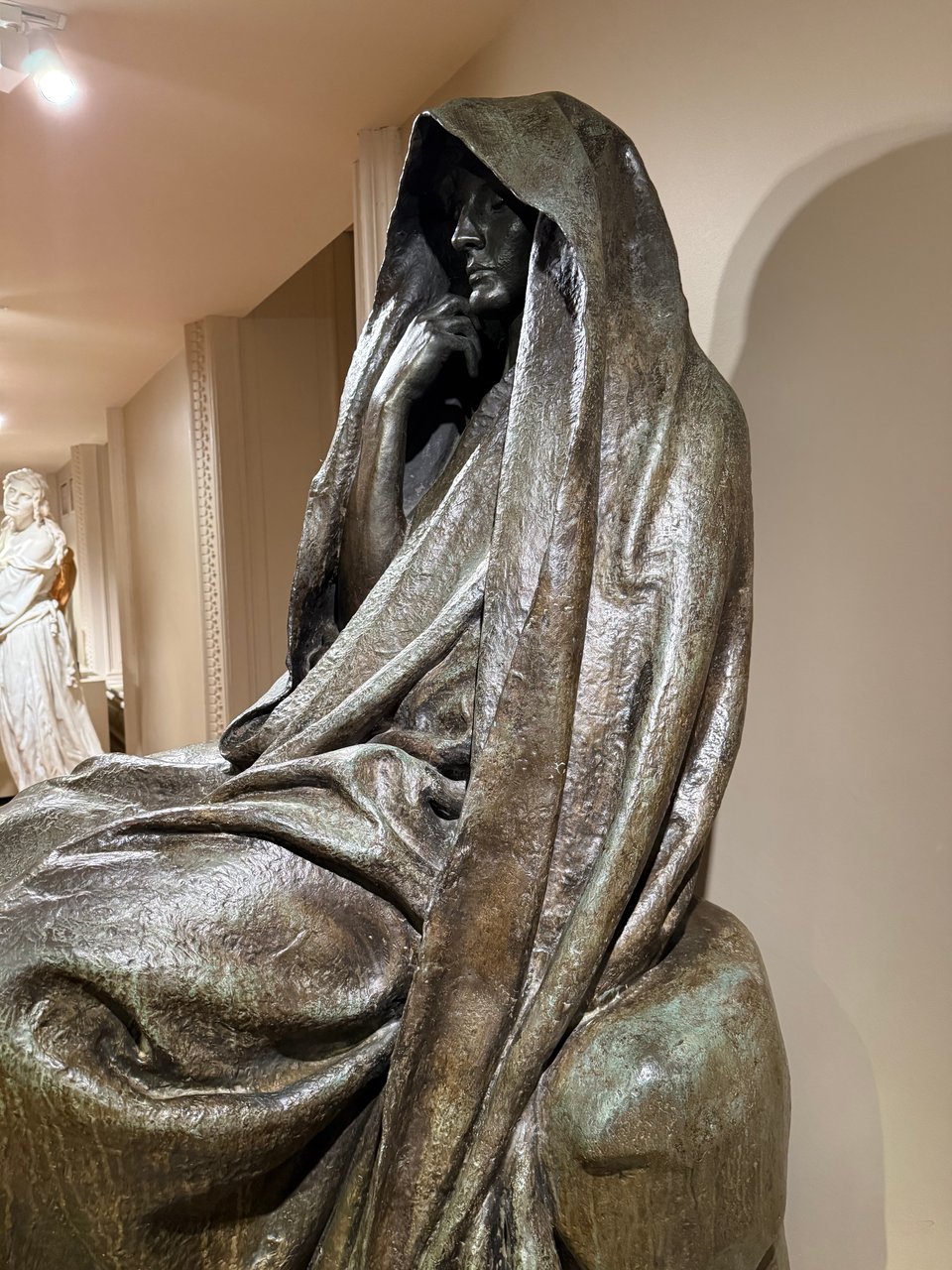 photo of bronze statue of a woman enshrouded in robes, one hand turned by her chin; her face is hooded
