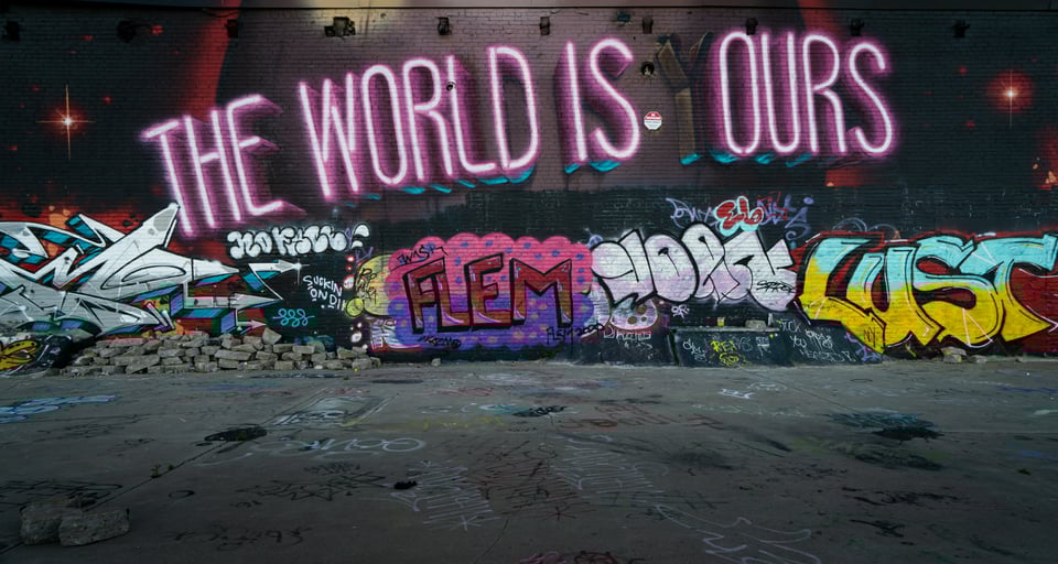 Pink neon lights spell out "The World Is Ours" over a graffitied wall. There is a burned out Y in front of the Ours, indicating the original message was "The World Is Yours"