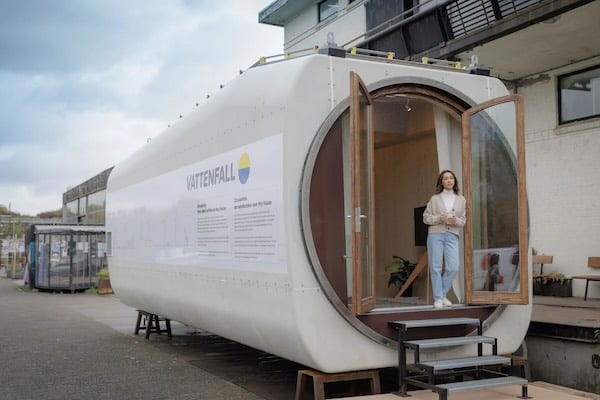 Perspectiva exterior de la antigua turbina de un aerogenerador convertida en una casa. Tiene una escalerita de cuatro peldaños apra entrar y puertas simples de madera y cristal. EL compartimento tiene el tamaño aproximado de la caja de un camión mediano. Hay una chica apoyada en la puerta.