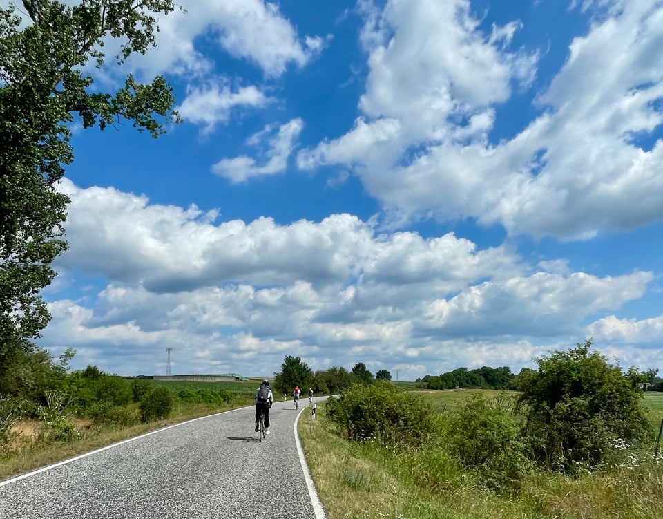 An empty curved road with some cyclists on it, the road is flanked by farm-land. In the background is expansive blue sky with lots of white clouds