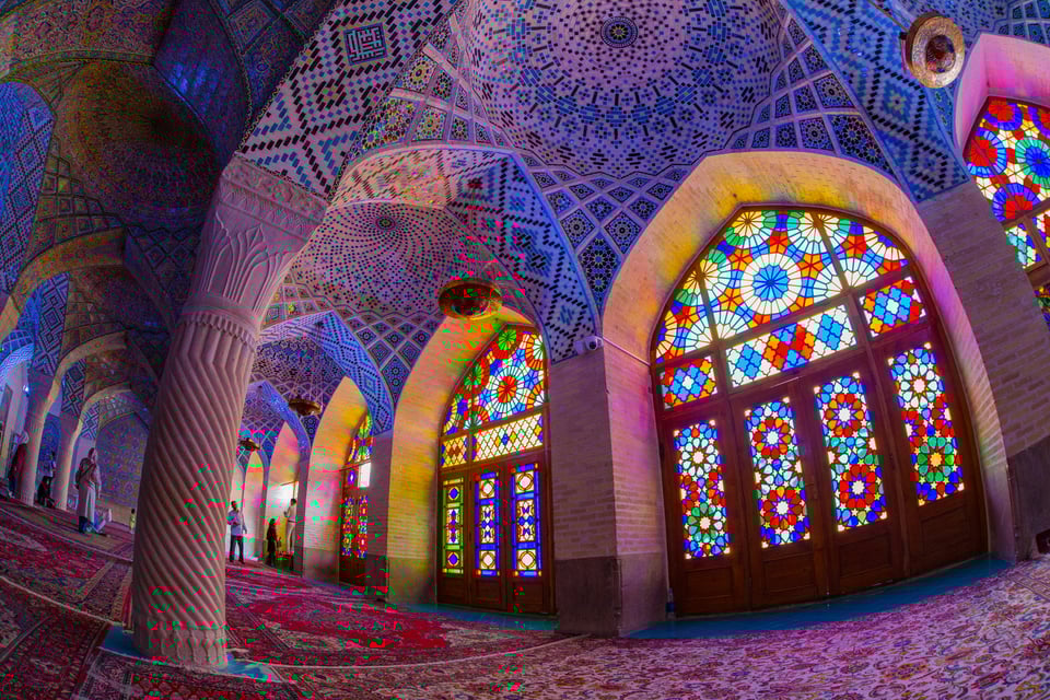 Nasir al-Mulk Mosque in Iran. Stained glass windows back lit by the sun, illuminate intricate tile work across a series of domes and arches, with similarly intricate rugs on the floor.