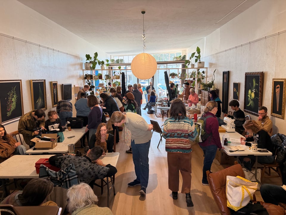 A small store-front, viewed from the back, showing six white fold-out tables, three on each side. On the left side people are fixing electronic appliances, and on the right side are mending projects. In the background are some plants and still more people. The author walks down the path between the tables.