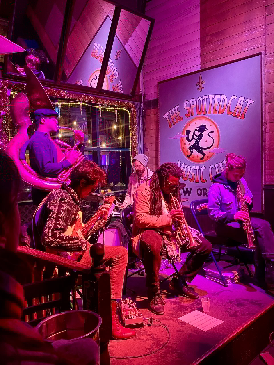 A small stage bathed in red-purple light, with five musicians deep in concentration. A sign for The Spotted Cat appears in the background.