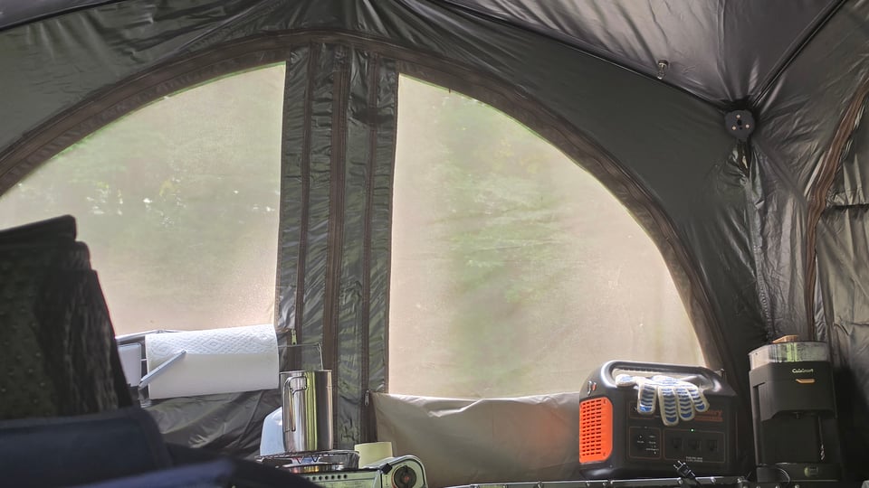 Interior view of a camping tent with large mesh windows looking out onto greenery, where a counter along the bottom holds a roll of paper towels, a stainless steel thermos, an orange-and-black portable power station, and a coffee maker.