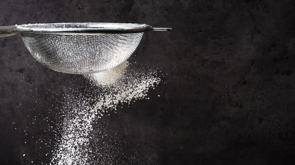 A photograph of flour falling through a sieve against a black background.