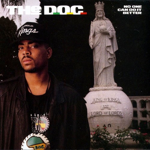 A color photograph of The D.O.C., a young Black man wearing a leather jacket, a black Sacramento Kings cap, and two medallions, possibly made of leather, showing Africa. He has a wary, suspicious look on his face. He is outside, possibly in a park somewhere. Behind him is a statue of I'm guessing Jesus, but wearing a crown, holding an orb, and a staff in the other hand. He's is on an orb-shaped pedastal that reads "KING OF KINGS AND LORD OF LORDS."