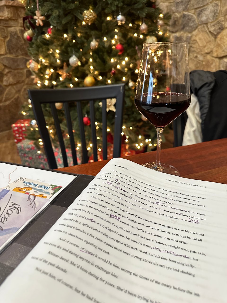 Cass's manuscript in a binder, open on a table in a winery in front of a glass of petit verdot, with a Christmas tree in the background.