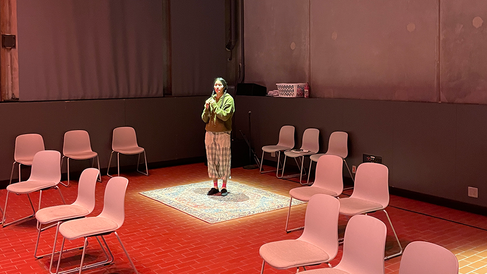 A woman stands in a pool of light with empty chairs around ready to welcome audience members.