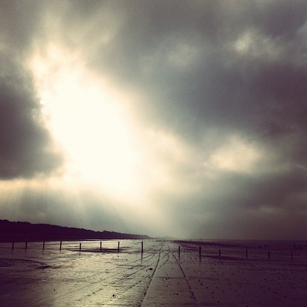 A wide, empty beach under heavy, overcast skies. A dramatic burst of light breaks through the thick clouds.
