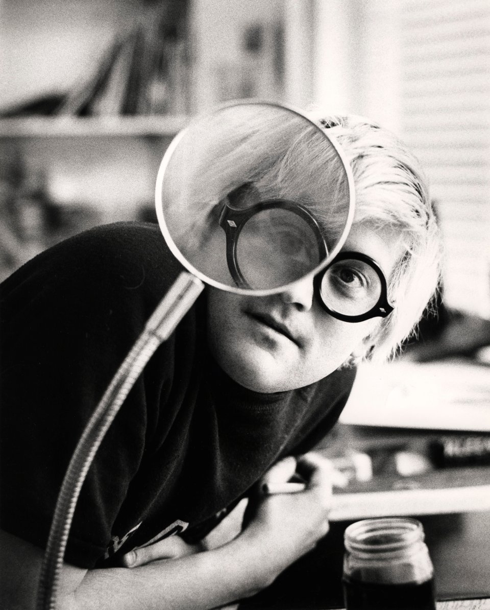 Black-and-white photograph of a young David Hockney leaning in to peer through a large round magnifier on a gooseneck arm, one eye enlarged behind the lens.