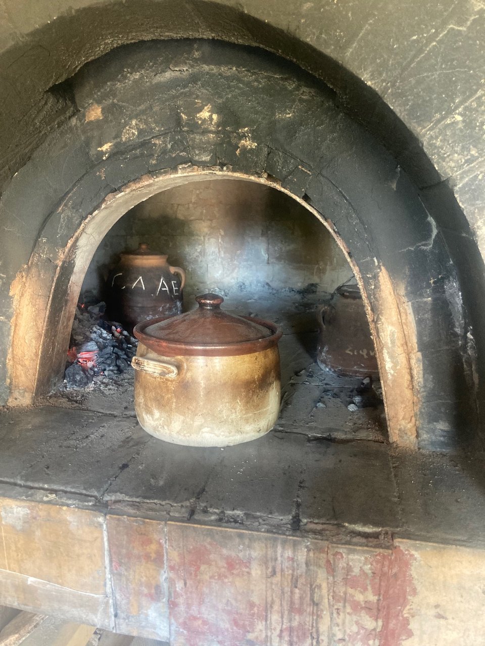 Round clay pot with a lid sitting at the opening arch of a wood-fired oven with other clay pots visible behind it.