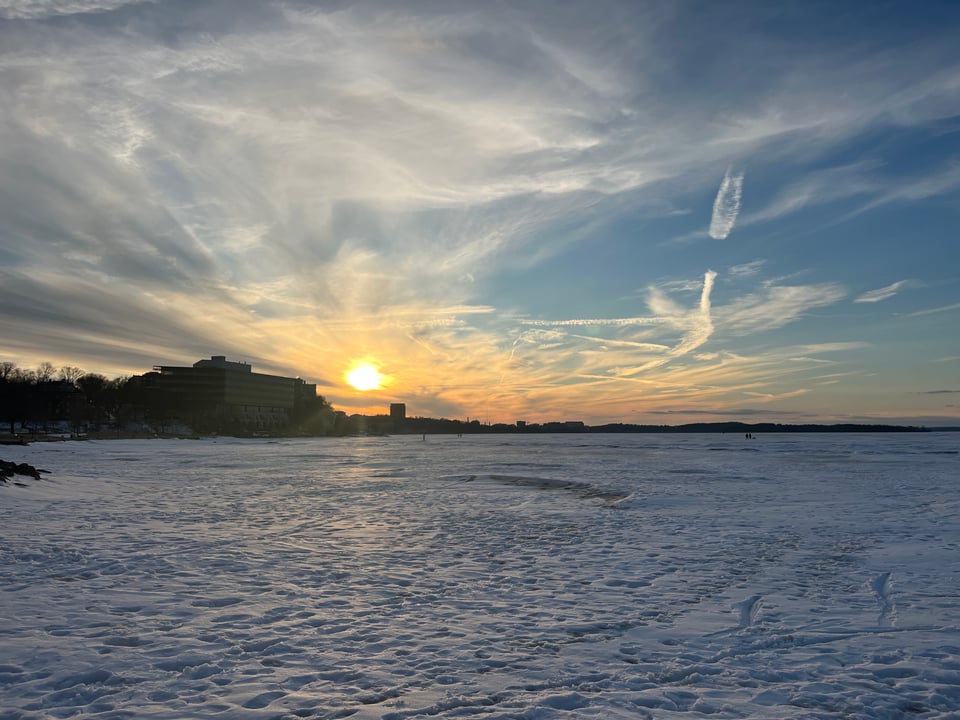 gold sun low in the sky, surrounded on two sides by ethereal clouds, over footprint-covered frozen lake