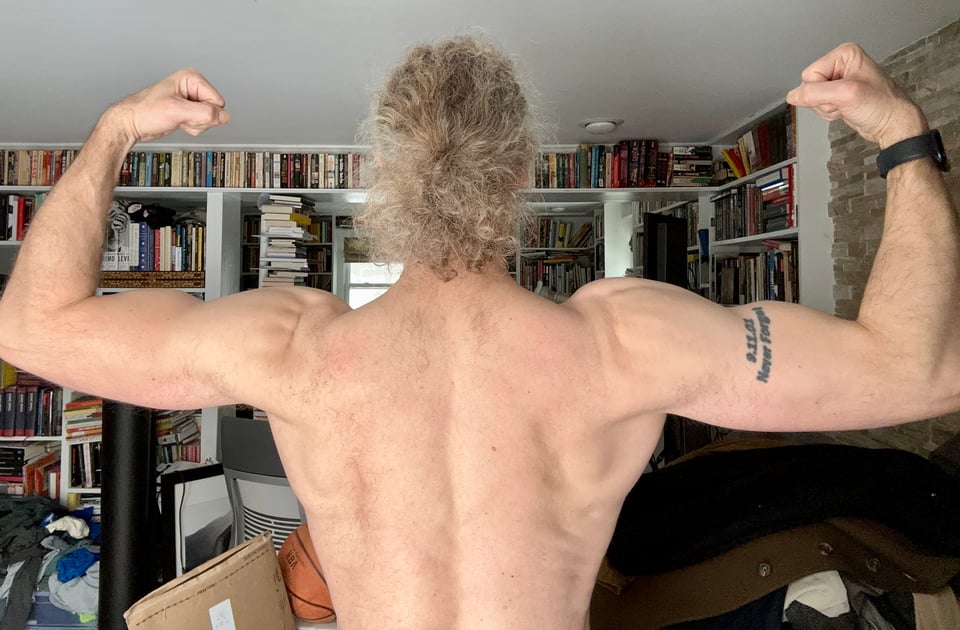 Photo of shirtless white man from behind, from the torso up, flexing his biceps. He has long curly hair tied back in a ponytail, and his shoulders & back look pretty good
