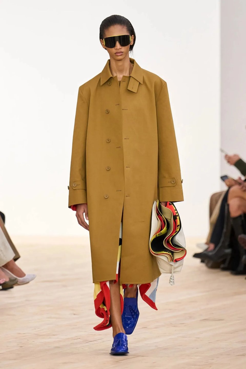 Loewe model in a very long tan overcoat that covers most but not all of a very colorful dress, the colorful ruffled edge of which emerges from beneath the bottom of the coat in a profusion of primary colors, a bit like a matador's cape. The look is paired with bright royal blue flat shoes and a big white Loewe bag that's open inside to reveal a bulbous red/yellow lining that matches the red/yellow lining of the dress. The model is wearing the same black sunglasses as in the previous look. It's all so good.