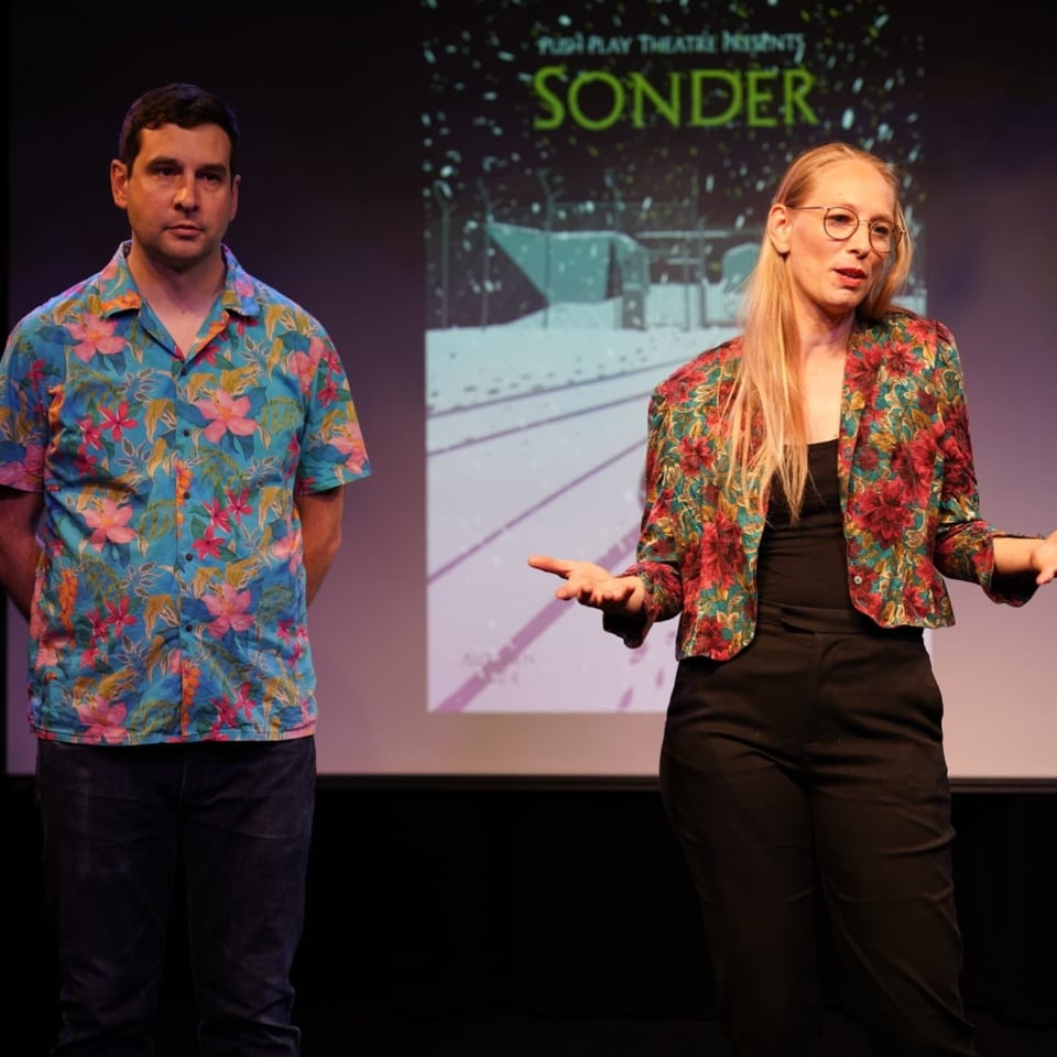 You see the projection of the show poster "Sonder" in the background with two people standing in front of it. One of them is saying something.