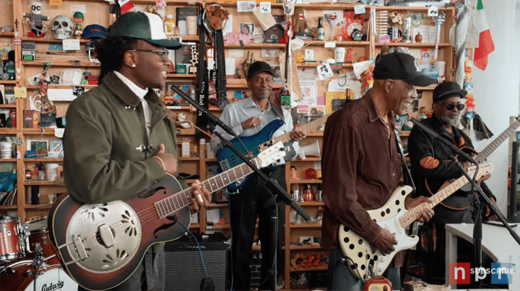 Miles Caton and Buddy Guy, each with their guitar, stand in front of microphones with other bandmates surrounding them in the NPR Tiny Desk studio.
