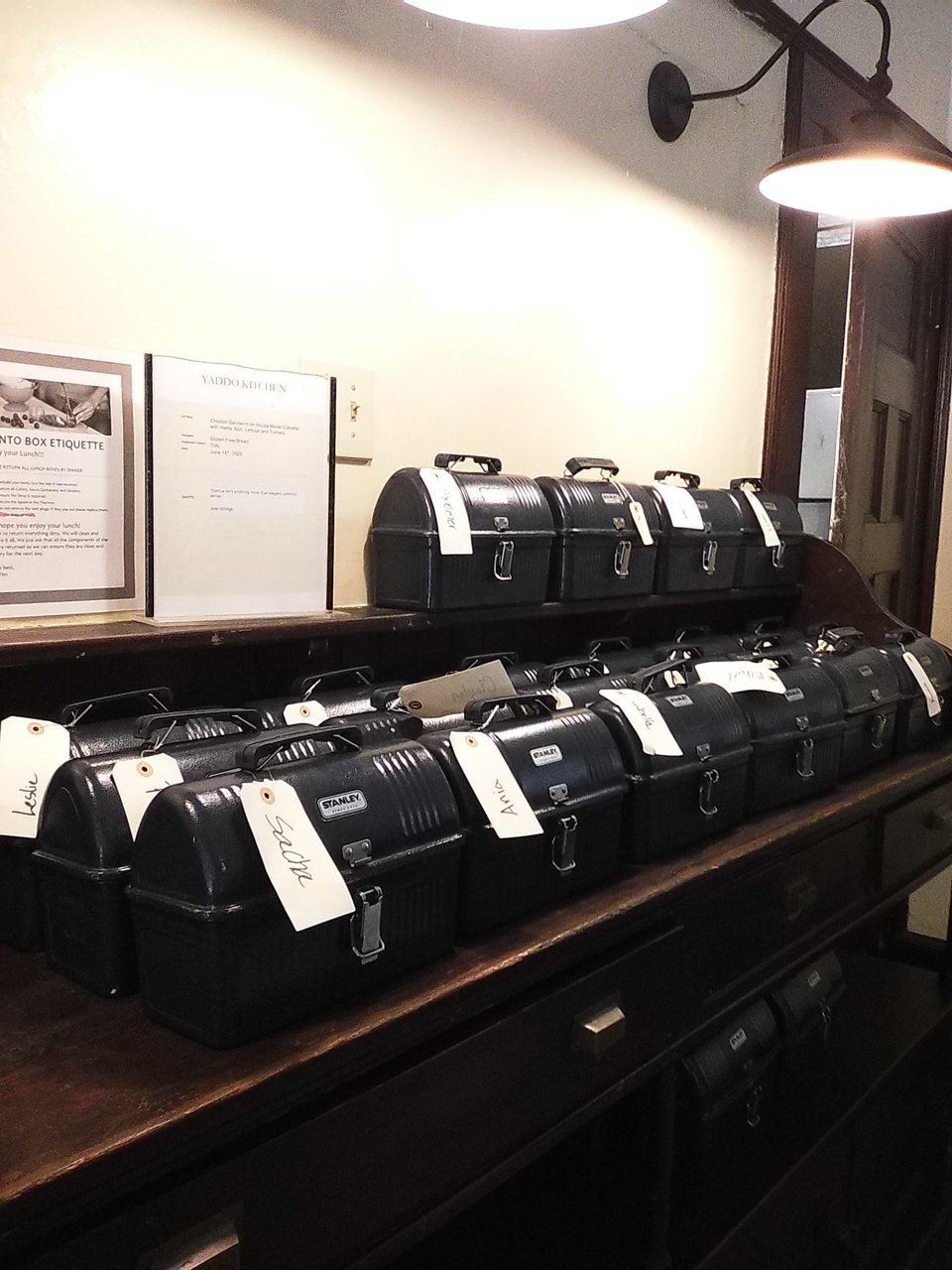 digital Instax photo of a series of black lunchboxes wit name tags on them, on a wooden table