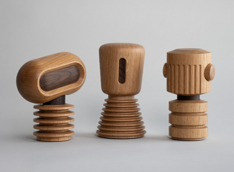 Three small wooden sculptures stand in a row against a neutral background, each shaped like a minimalist robot. Their forms combine smooth, rounded tops with ribbed or grooved cylindrical bases, showcasing light oak and darker walnut accents. They've got a warm, handcrafted aesthetic, with a playful look