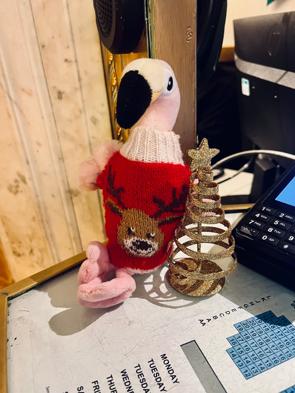 A small pink toy flamingo sitting on a desk, with a red knitted Christmas jumper that has a picture of a reindeer on the front.