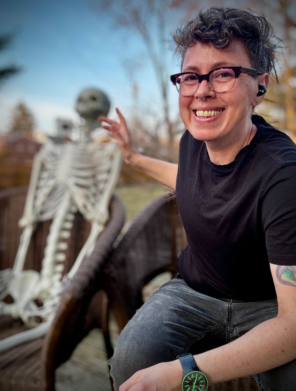 A white man sits in a chair, leaning forward. He is wearing a black t-shirt and grey jeans, smiling at the camera. Grey curly hair piled high on his head. He is gesturing vaguely at a plastic Halloween skeleton decoration sitting in a similar chair, though the plastic skeleton is out of focus.