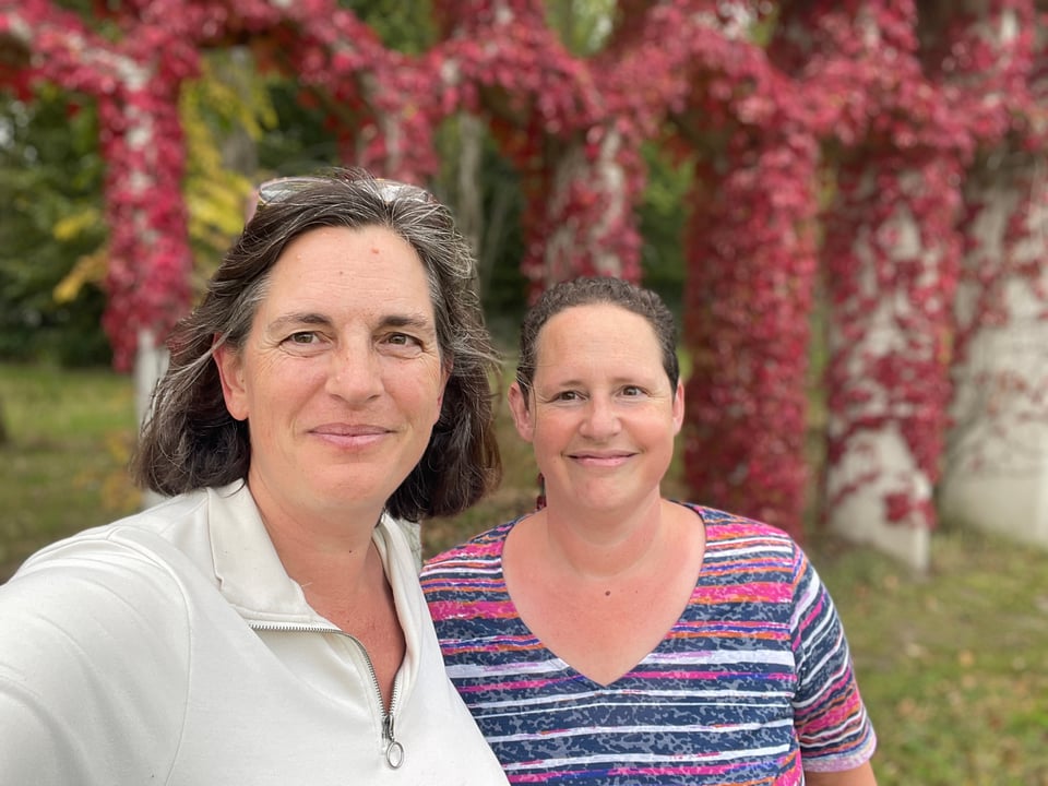 Two women smile at the camera and behind them is a "honeycomb" structure covered in red ivy.