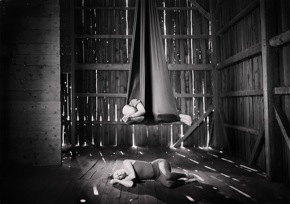 black and white, very crisply focused image of the old barn interior, light coming between the boards. Nicole is curled up partly-fetal on the floorboards, and I am mimicking her position directly above in the aerial sling.