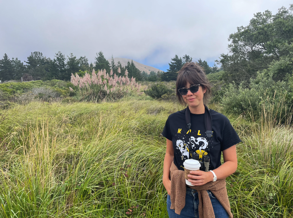 Jenny Odell wearing a KALX radio T-shirt, binoculars, and sunglasses, holding a coffee, standing in front of a coastal meadow with morning fog just beginning to lift