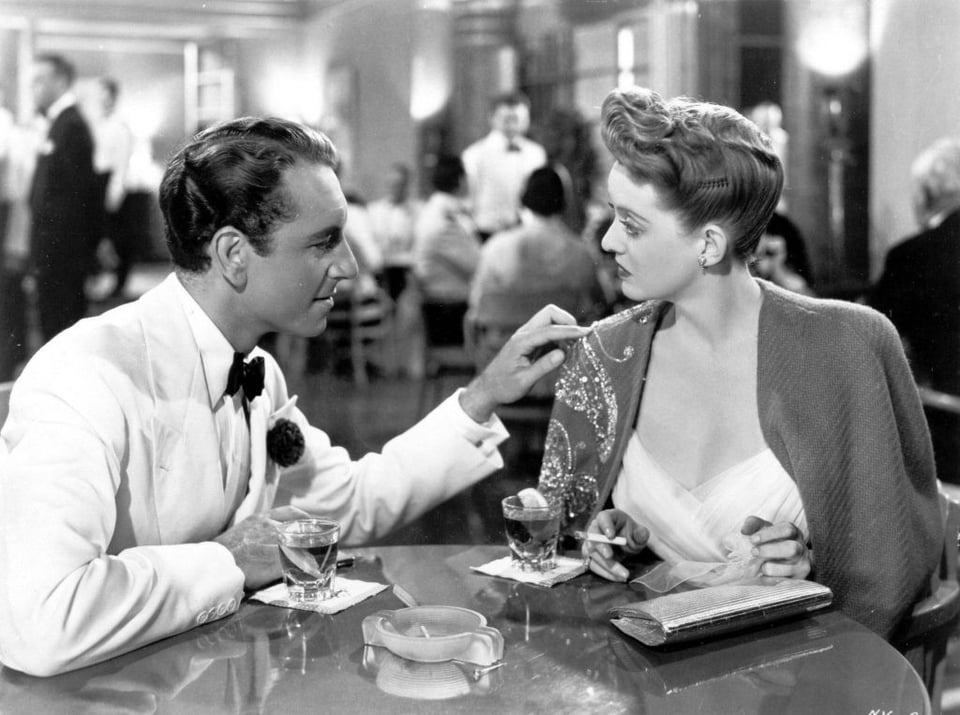 Jerry (Paul Henreid) and Charlotte (Bette Davis) in "Now, Voyager" (1942) sitting at a table in evening wear. Jerry is lightly touching the embroidery on the shoulder of Charlotte's cape.