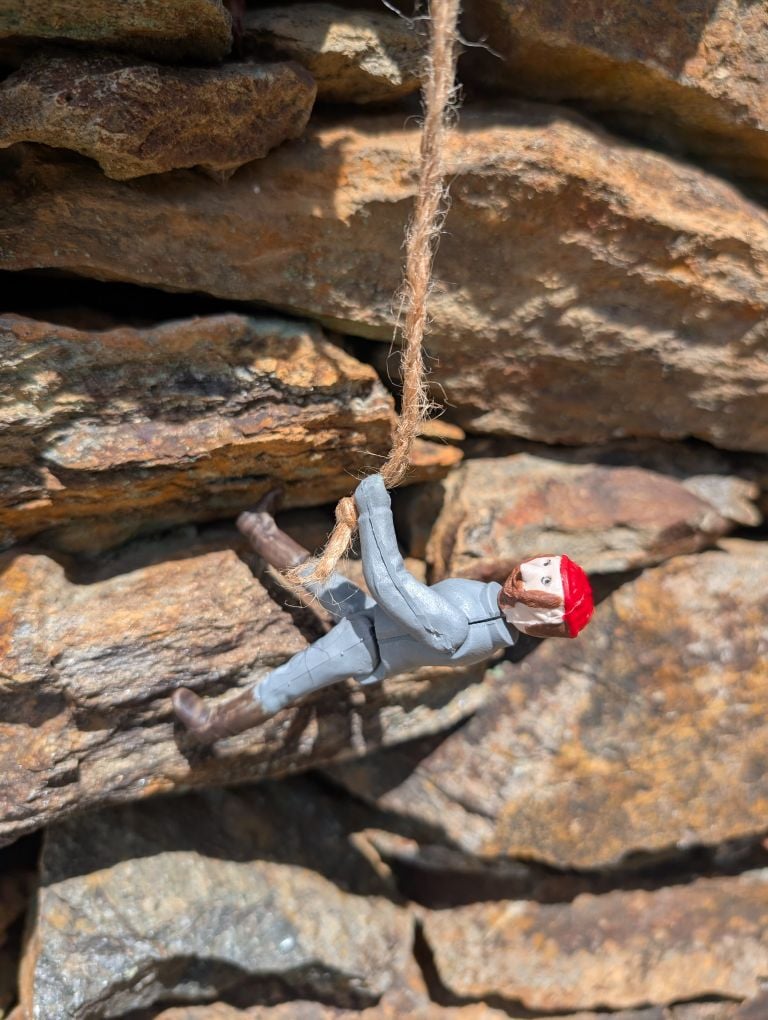 One of our injection molded toys, a man in a grey suit with boots and a red beanie, climbs a rock.