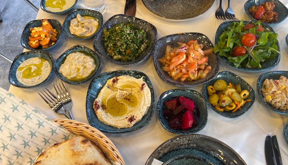 A table spread with at least 13 small dark blue ceramic dishes filled with different salads, plus a basket of bread