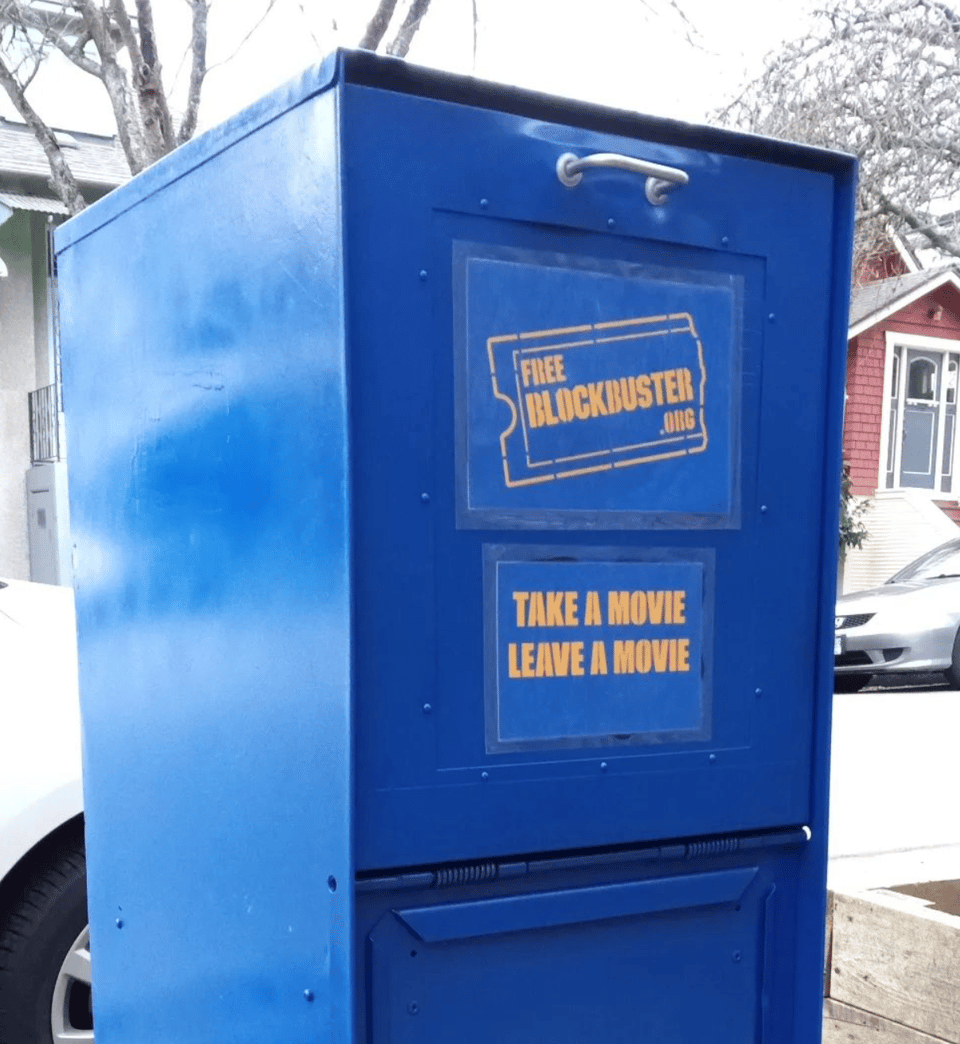 A repurposed newspaper box painted in bright blockbuster blue. On the face is a sign in yellow that says "Free Blockbuster.org" and "Take a Movie, Leave a Movie." Behind the box, you can see some details of parked cars and houses.