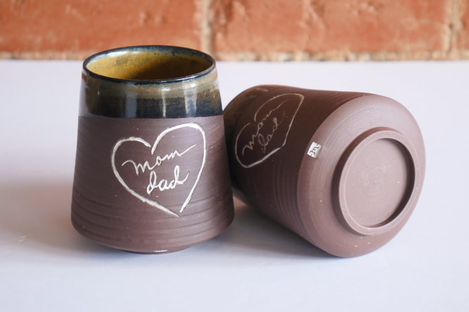 Two ceramic tumblers made from a rich brown clay, one of them on it's side to show the base. The body curves up and out from an inset foot ring, then suddenly angles in at a gentle slope towards the rim. There are four neatly spaced rings carved near the base, and then a hand-drawn heart inlaid with white, and the words mom and dad in two different handwritings. The rims are glazed black with floating organic patches of gold.