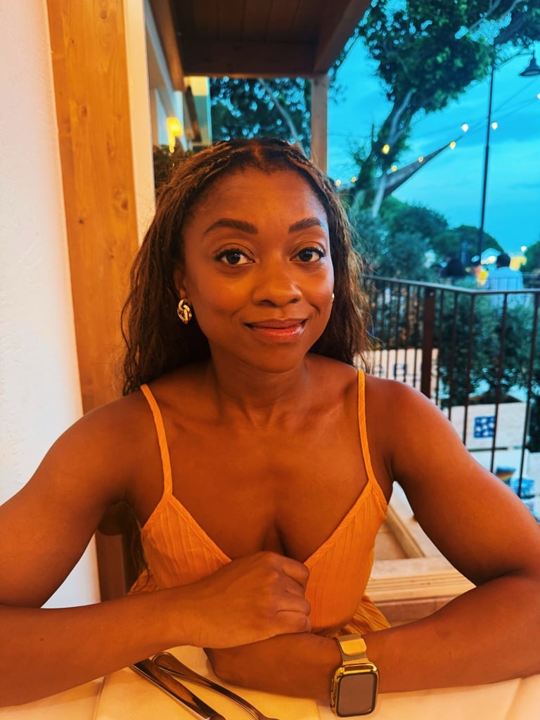 Me in an orange spaghetti-strap dress sitting at a restaurant table, smiling at the camera. I'm wearing gold earrings and a gold-banded smartwatch, with trees and evening lights visible outside behind me.