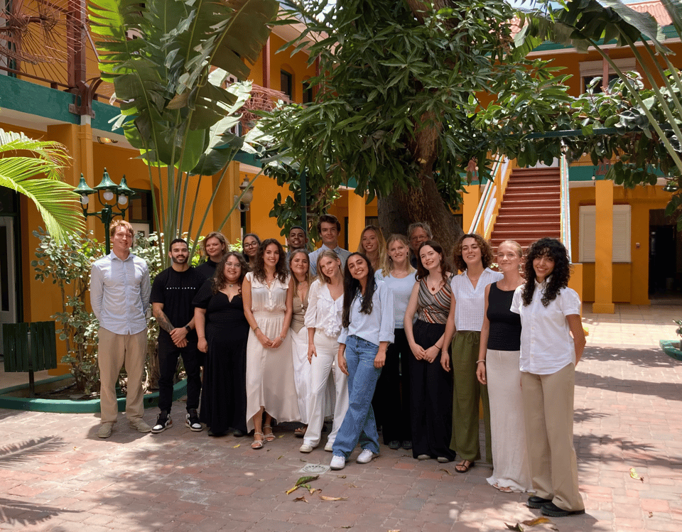 A picture of the UACU students standing in the court yard of the Marie de la Salle building of the University of Aruba. They are all smiling happily into the camera, standing in front of a huge mango tree.