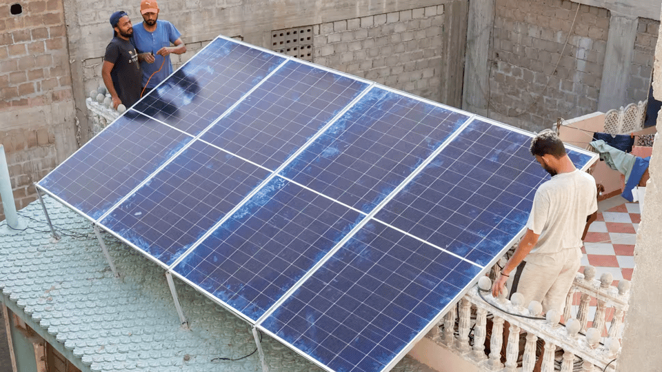 A photo of three Pakistani men standing around a large solar panel installed on the deck of an old-fashioned brick building. There is a interesting aesthetic difference between the age of the building and the high-tech look of the shining black solar panels.