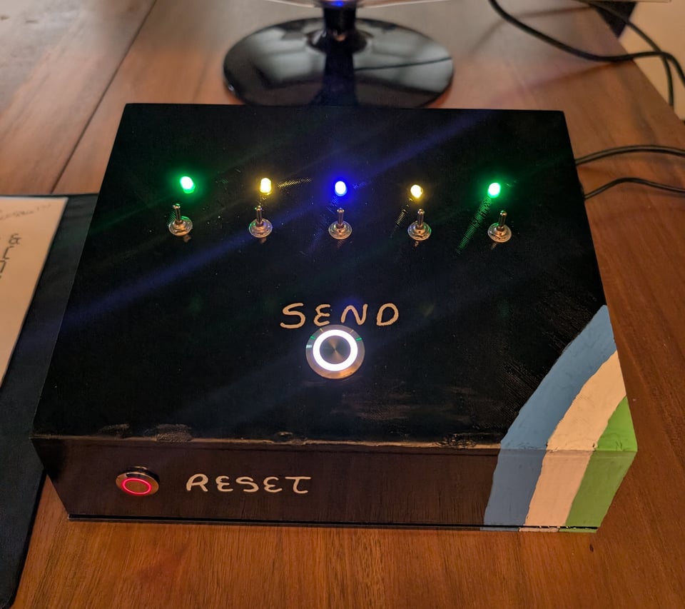 A photo of the NLB typing console: a black box with 5 toggle switches, a big metal SEND button, and a smaller red RESET button on the side