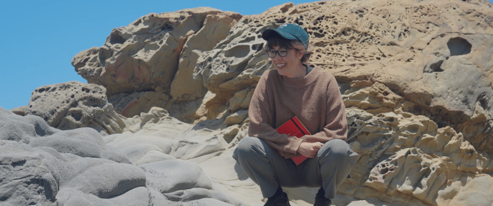 Jenny Odell wearing a green baseball cap and brown sweater, holding a red notebook and perched on some sandstone showing coastal erosion