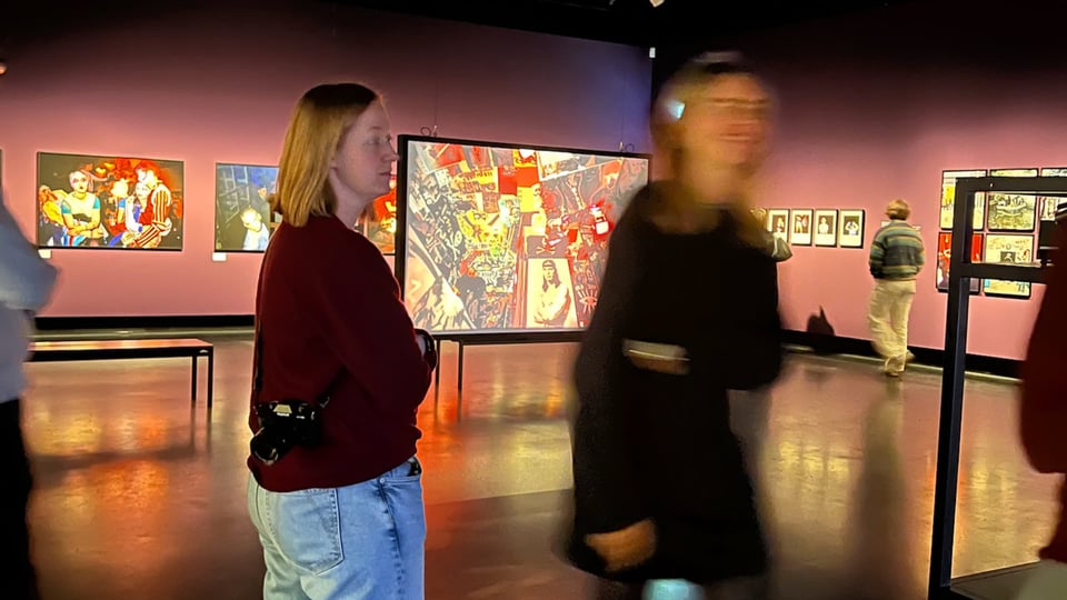 Photo of two people standing in an art gallery with large photo prints on the walls, dimly lit, one person blurry in motion