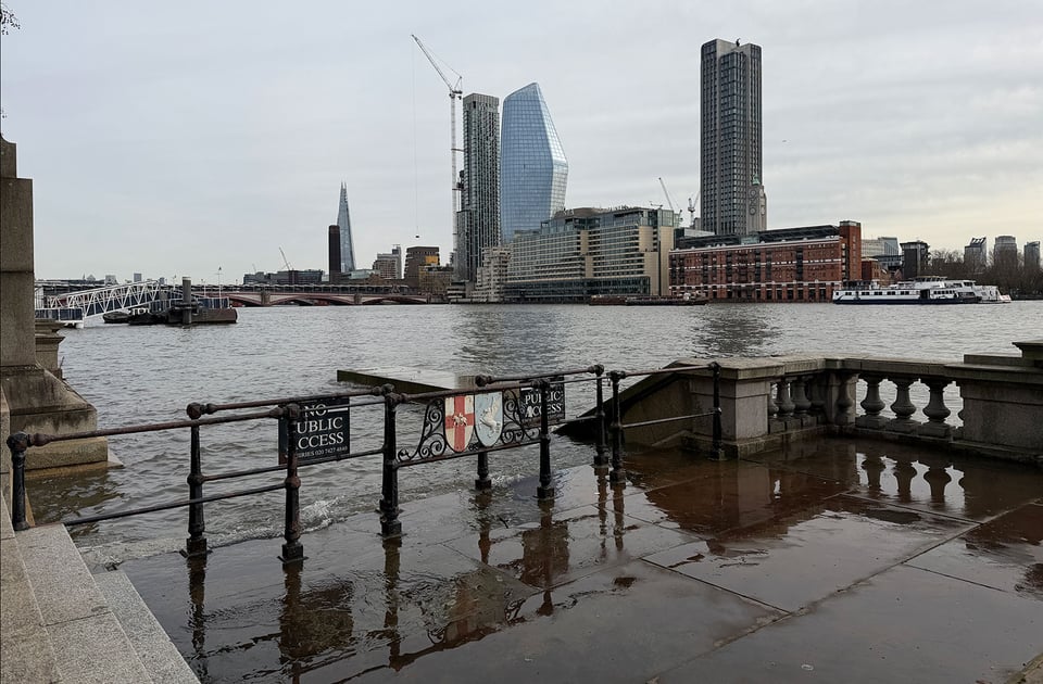 Thames river in London at high tide is lapping at the top of a staircase that could give access to boats. Skyscrapers on the opposite bank are seen in the distance.