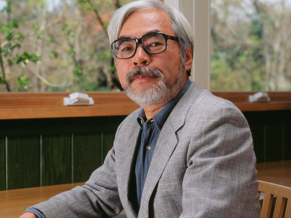 An image of Japanese Anime director Hayao Miyazaki, with close cropped gray hair and trimmed beard and black glasses, seated at a wooden table while wearing a gray suit with blue undershirt