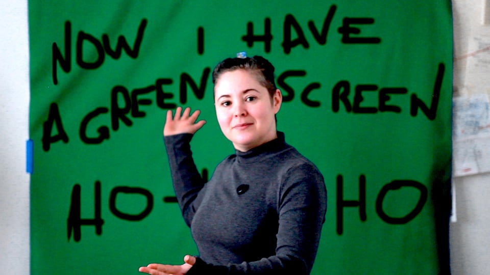 Sparks gestures to a green-screen fabric on which is written in block hand-lettering 'NOW I HAVE A GREEN SCREEN HO-HO-HO'