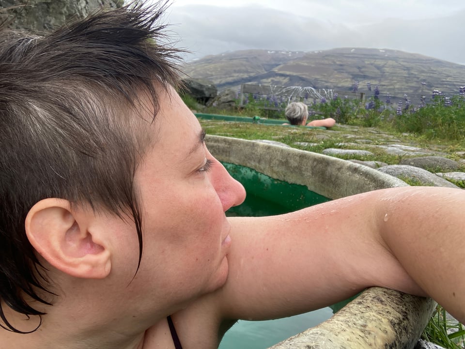 Me, a white person with short brown wet hair, leaning on the edge of a geothermal hot tub looking out into the distance
