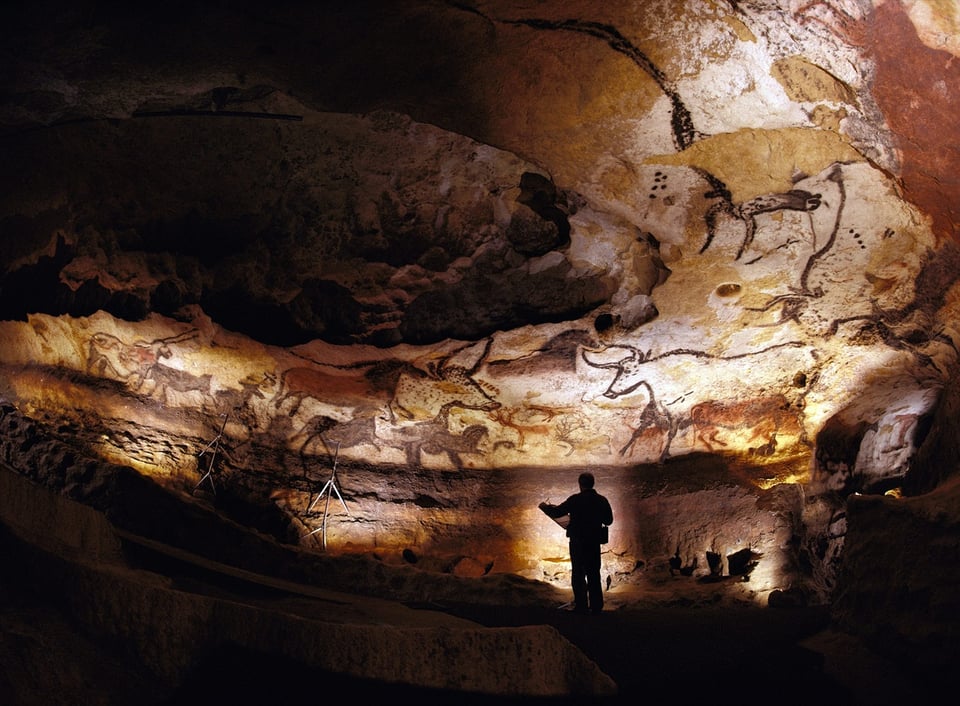 A photo of a man standing in a large cave covered with cave art. The art is large animals with horns. It's dark and there are moody lights illuminating the walls; the man is a shadow