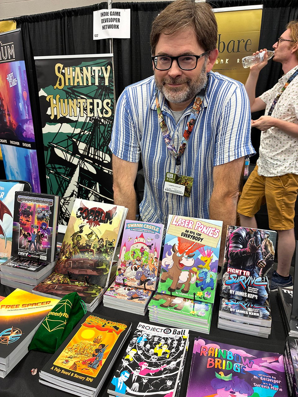 A picture of me, not just my hand this time, behind a table loaded up with RPGs for sale. Some of my own are in front of me, like Laser Ponies, Swann Castle, and Project 8Ball.